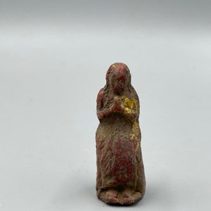 Puede incluir: Una pequeña figura de arcilla roja de una mujer con detalles dorados. La figura está sentada con las manos juntas frente a ella.