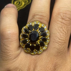Może przedstawiać: Złoty pierścionek z dużym, owalnym czarnym kamieniem pośrodku, otoczonym mniejszymi, czarnymi kamieniami w kształcie płatków i przezroczystymi kamieniami akcentującymi. Pierścionek ma kwiatowy wzór.