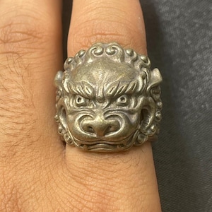 Könnte beinhalten: Ein silberfarbener Ring mit einem detaillierten Löwenkopf-Design. Der Ring zeigt aufwendige Schnitzereien der Löwenmähne, der Gesichtszüge und der Gesamtform. Der Ring wird an einem Finger getragen, was seine Größe und Handwerkskunst hervorhebt.