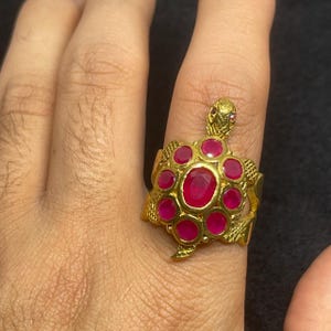 Może przedstawiać: Złoty pierścionek w kształcie żółwia z centralnym owalnym rubinem otoczonym mniejszymi okrągłymi rubinami. Głowa i nogi żółwia są szczegółowe. Pierścionek jest noszony na palcu.