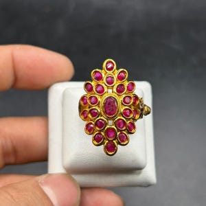 Może przedstawiać: Złoty pierścionek z czerwonymi kamieniami szlachetnymi. Pierścionek ma owalny kamień centralny otoczony mniejszymi, okrągłymi kamieniami. Pierścionek jest prezentowany na białej powierzchni. Projekt przypomina kompozycję kwiatową.