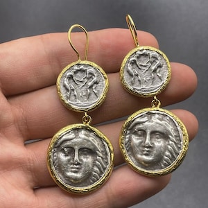 May include: A pair of gold and silver dangle earrings featuring two round coin-shaped charms. The top charm features a silver design of three figures, while the bottom charm features a silver design of a woman's face.