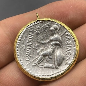 May include: A silver coin pendant with a gold rim. The coin features a depiction of a woman holding a spear and shield. The inscription on the coin reads "AEMILIA", "PAETINA", and "COS".