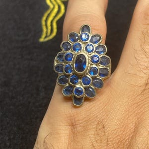 Może przedstawiać: Srebrny pierścionek z owalnym kamieniem centralnym i otaczającymi niebieskimi kamieniami szlachetnymi. Pierścionek ma kwiatowy lub klastrowy wzór, z niebieskimi kamieniami rozmieszczonymi wokół centralnego kamienia. Pierścionek jest noszony na palcu.