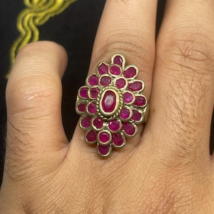 Może przedstawiać: Srebrny pierścionek z kwiatowym wzorem, osadzony z licznymi owalnymi, głęboko czerwonymi kamieniami szlachetnymi. Centralny kamień otoczony jest złotą obwódką. Pierścionek jest prezentowany na tle w kolorze skóry.