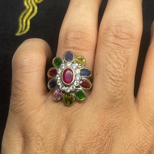 Może przedstawiać: Srebrny pierścionek z centralnym owalnym rubinem otoczonym małymi białymi kamieniami. Projekt pierścionka obejmuje kwiatowy układ kolorowych kamieni szlachetnych, w tym fioletowych, żółtych, niebieskich i zielonych, osadzonych na srebrnej obrączce.