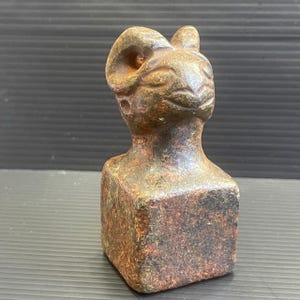 Puede incluir: Una pequeña escultura de piedra de color verde oscuro y marrón que representa la cabeza de un carnero con cuernos. La escultura está sobre una base cuadrada.