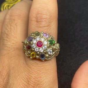 Może przedstawiać: Złoty pierścionek z centralnym czerwonym kamieniem szlachetnym, otoczonym białymi kamieniami i zewnętrznym okręgiem z kolorowych kamieni szlachetnych. Pierścionek ma bogaty, kwiatowy wzór.