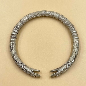 Puede incluir: Un brazalete de plata con intrincados diseños florales y de dragones. El brazalete está abierto y tiene un espacio en la parte superior.