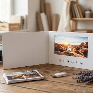 May include: A white video greeting card featuring a landscape screen displaying a desert sunset. The card has six circular control buttons below the screen. A stack of small photographs tied with twine and a white USB flash drive sit nearby.