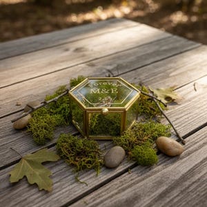 Puede incluir: Un joyero hexagonal de cristal con marco dorado con la inscripción "forever M&E 2.5" descansa sobre una superficie de madera, rodeado de musgo verde, pequeñas piedras y una hoja. La caja está cerrada y contiene un anillo.