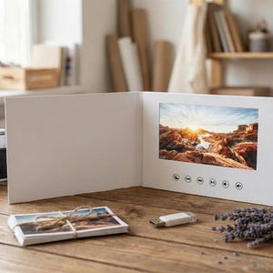 May include: A white video greeting card featuring a landscape screen displaying a desert sunset scene. The card has a series of circular control buttons below the screen. A small white USB flash drive is positioned on the wooden surface nearby.