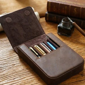Pode incluir: Um estojo de couro castanho escuro para canetas com tampa articulada, apresentando quatro compartimentos individuais contendo canetas com acabamentos met&aacute;licos em preto, dourado, cobre e azul. O interior &eacute; forrado com camur&ccedil;a castanha macia.