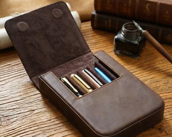 May include: A dark brown leather pen case with a hinged lid, featuring four individual slots containing pens with black, gold, copper, and blue metallic finishes. The interior is lined with soft brown suede, and the exterior has a smooth matte texture.