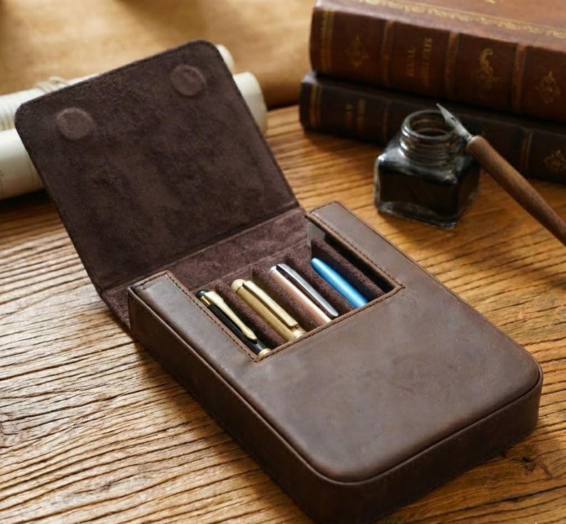Pode incluir: Um estojo de couro castanho escuro para canetas com tampa articulada, apresentando quatro compartimentos individuais contendo canetas com acabamentos met&aacute;licos em preto, dourado, cobre e azul. O interior &eacute; forrado com camur&ccedil;a castanha macia.