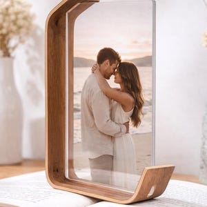 May include: A wooden photo frame with a curved design, holding a picture of a couple embracing on a beach. The frame is made of light brown wood and sits on an open book. The photo frame is approximately 8 inches tall.