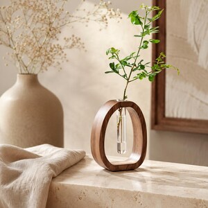 Puede incluir: Un jarr&oacute;n de madera de forma ovalada con un inserto de tubo de vidrio transparente que contiene un tallo de planta verde. El jarr&oacute;n es de madera oscura y se encuentra sobre una superficie de piedra de color claro. Un jarr&oacute;n beige con flores secas en el fondo.