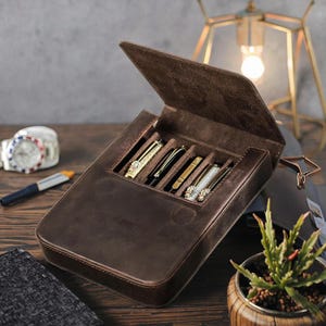 Pode incluir: Um estojo de armazenamento em couro castanho escuro com tampa articulada, concebido para guardar quatro instrumentos de escrita. O interior apresenta quatro ranhuras individuais contendo canetas com acabamentos met&aacute;licos, texturizados e decorativos. O estojo tem uma textura macia semelhante a camur&ccedil;a e uma forma retangular com cantos arredondados.