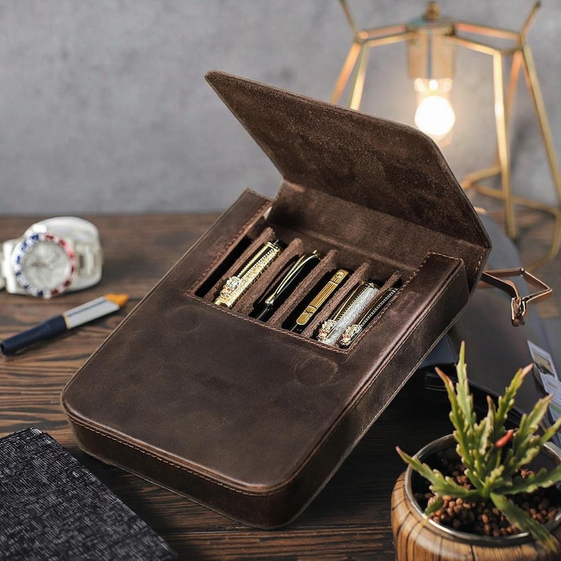 Pode incluir: Um estojo de armazenamento em couro castanho escuro com tampa articulada, concebido para guardar quatro instrumentos de escrita. O interior apresenta quatro ranhuras individuais contendo canetas com acabamentos met&aacute;licos, texturizados e decorativos. O estojo tem uma textura macia semelhante a camur&ccedil;a e uma forma retangular com cantos arredondados.