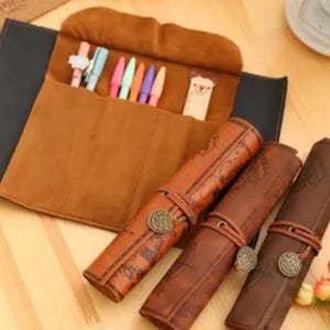 May include: A brown leather pen roll with a map design, featuring a flap with pens and a bookmark. The roll is secured with a leather strap and a decorative metal button. The open roll reveals a brown suede interior.