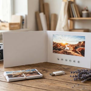 May include: A white video greeting card showing a landscape image with a sunset. A stack of photos tied with twine, a USB drive, a vintage camera, and lavender sprigs are on a wooden surface.