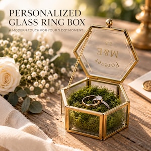 Peut inclure: Une boîte à bagues hexagonale en verre avec un cadre en métal doré et un couvercle à charnière. Le couvercle porte l'inscription M&E et FOREVER. L'intérieur contient de la mousse verte et deux bagues. Une chaînette dorée relie le couvercle.