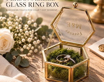 May include: A hexagonal glass ring box with a gold metal frame and a hinged lid. The lid features the text M&E and FOREVER. Inside, the box contains green moss and two rings. A gold chain connects the lid to the base of the container.