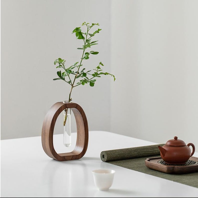 Puede incluir: Un jarr&oacute;n de madera moderno, de forma ovalada, con un inserto de tubo de vidrio que contiene un tallo verde y frondoso. El jarr&oacute;n es de color marr&oacute;n intenso y se encuentra sobre una superficie blanca. Tambi&eacute;n se ven una peque&ntilde;a taza de t&eacute; blanca y una tetera marr&oacute;n.