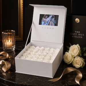 Könnte beinhalten: Eine weiße Geschenkbox mit eingebautem Bildschirm, der ein Video zeigt, gefüllt mit weißen Rosen. Die Box ist geöffnet und zeigt die Blumen und den Bildschirm. Der Text "THE GIFT BOX ATELIER" steht auf einer schwarzen Box.