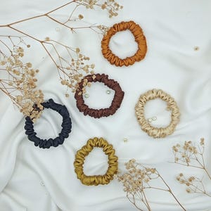 May include: An assortment of five silk scrunchies in various colors: black, brown, gold, tan, and rust. The scrunchies are arranged on a white satin surface, with dried flowers and small pearls scattered around them.
