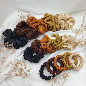 May include: An assortment of satin scrunchies in various colors, including black, brown, gold, and olive green, are arranged on a white fabric. Some scrunchies are bunched, while others are in a ring shape. The scrunchies have a shiny, smooth texture.
