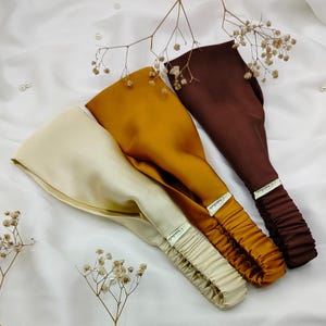 May include: Three satin headbands in cream, mustard yellow, and brown. Each headband features a wide, ruched elastic band and a small label. The headbands are arranged on a white, textured surface with small floral accents.
