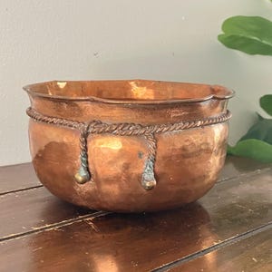 May include: A copper-colored bowl with a decorative rope design around the middle. The bowl has a rounded shape with a slightly scalloped rim. Two rope tassels with brass-colored accents hang from the rope design.