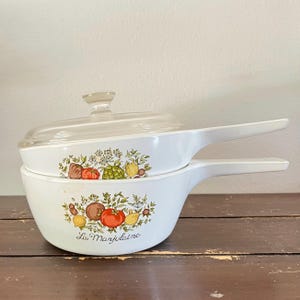 May include: Two white, handled saucepans with a clear glass lid. Each pan features a colorful illustration of vegetables and herbs. The text "La Marjolaine" is printed on the front of the bottom pan. The pans are stacked on top of each other.