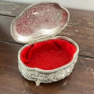 Puede incluir: Una caja de alhajas de plata con un diseño ornamentado y tres patas. La caja está abierta y forrada con terciopelo rojo.