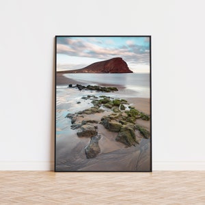 Può includere: Una fotografia incorniciata di una formazione rocciosa rossa su una spiaggia con rocce ricoperte di muschio verde in primo piano e un cielo blu con nuvole sullo sfondo.