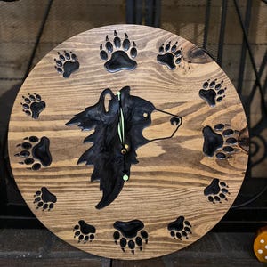 May include: A wooden clock with a black bear head silhouette and paw prints around the edge. The clock face is made of wood with a dark brown stain.