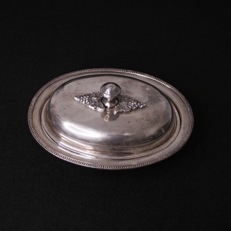 May include: A silver oval-shaped butter dish with a lid. The lid has a knob with a grapevine design.