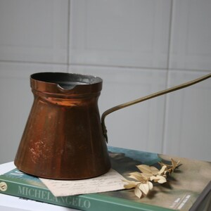 Pode incluir: Uma cafeteira de cobre com uma alça de latão. A cafeteira tem uma forma cónica. Repousa sobre um livro com a palavra "MICHELANGELO" visível. Um pequeno pedaço de papel e folhas secas também estão presentes. Cerca de 15 cm de altura.