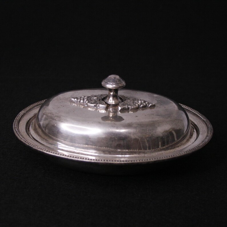 May include: Silver oval butter dish with a lid and a decorative knob. The lid has a floral design.