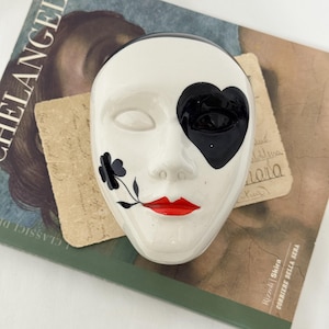 Peut inclure: Un masque en céramique blanc avec un cache-œil noir en forme de cœur, des lèvres rouges et un motif floral noir. Le masque repose sur un livre avec le mot "MICHELANGELO" visible.