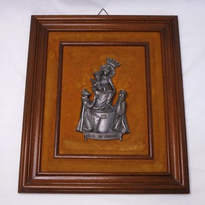 Puede incluir: Una escultura en relieve de metal enmarcada que representa una figura coronada sosteniendo a un niño, flanqueada por dos figuras. El relieve está sobre un fondo marrón claro dentro de un marco de madera marrón oscuro. La escultura tiene el texto "AVE MARIA" y "L.V. DI PONTE".