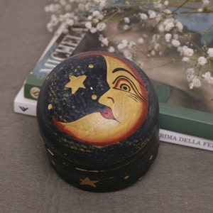 Könnte beinhalten: Eine runde Holzkiste mit handbemaltem Design eines Halbmondes und eines Gesichts. Der Mond ist gelb mit roten Lippen und einem Auge, und der Hintergrund ist dunkelblau mit weißen Sternen.
