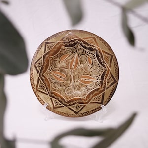 Peut inclure: Une assiette décorative ronde en bois avec des motifs floraux et géométriques complexes. L'assiette présente un motif floral central dans des tons de marron et d'or, entouré d'une bordure détaillée. L'assiette est présentée sur un support transparent.