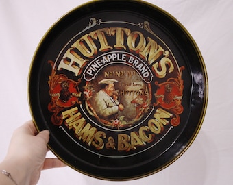 Può includere: Un vassoio da portata vintage in metallo con sfondo nero e lettere dorate. Il vassoio presenta un logo per Hutton's Pine-Apple Brand Hams & Bacon. Il logo include un uomo con un cappello da cuoco e un ananas.