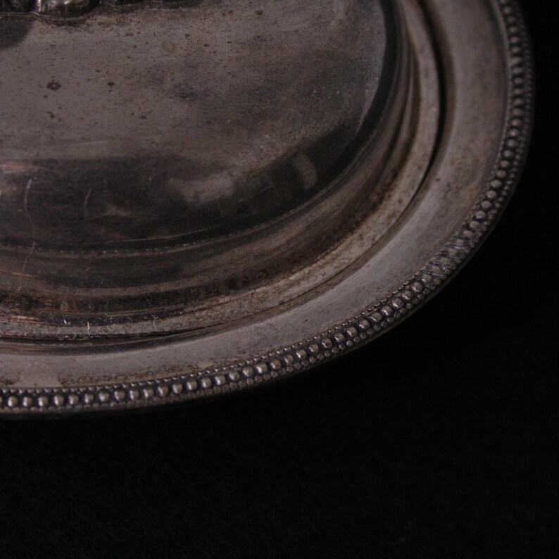 May include: Close-up of a silver butter dish with a beaded edge. The lid is slightly ajar, revealing the smooth, silver interior.