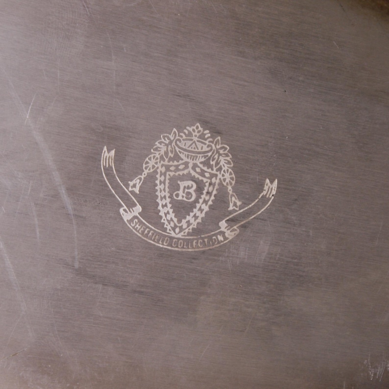 May include: A silver serving tray with an engraved crest featuring a shield with the letter "B" inside, surrounded by a wreath and a banner that reads "Sheffield Collection".