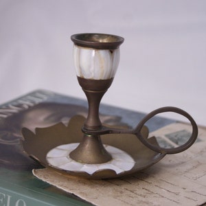 May include: A brass candlestick with a white mother-of-pearl inlay. The candlestick has a round base with a scalloped edge and a handle.
