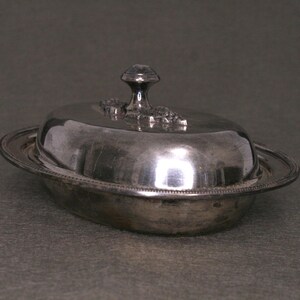May include: A silver butter dish with a domed lid and a decorative knob. The dish has a beaded edge and a smooth, reflective surface.