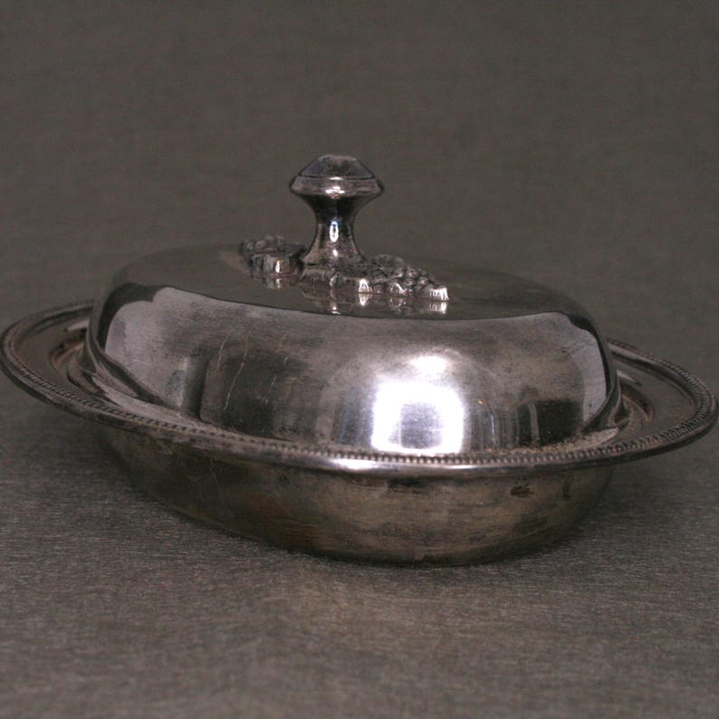 May include: A silver butter dish with a domed lid and a decorative knob. The dish has a beaded edge and a smooth, reflective surface.